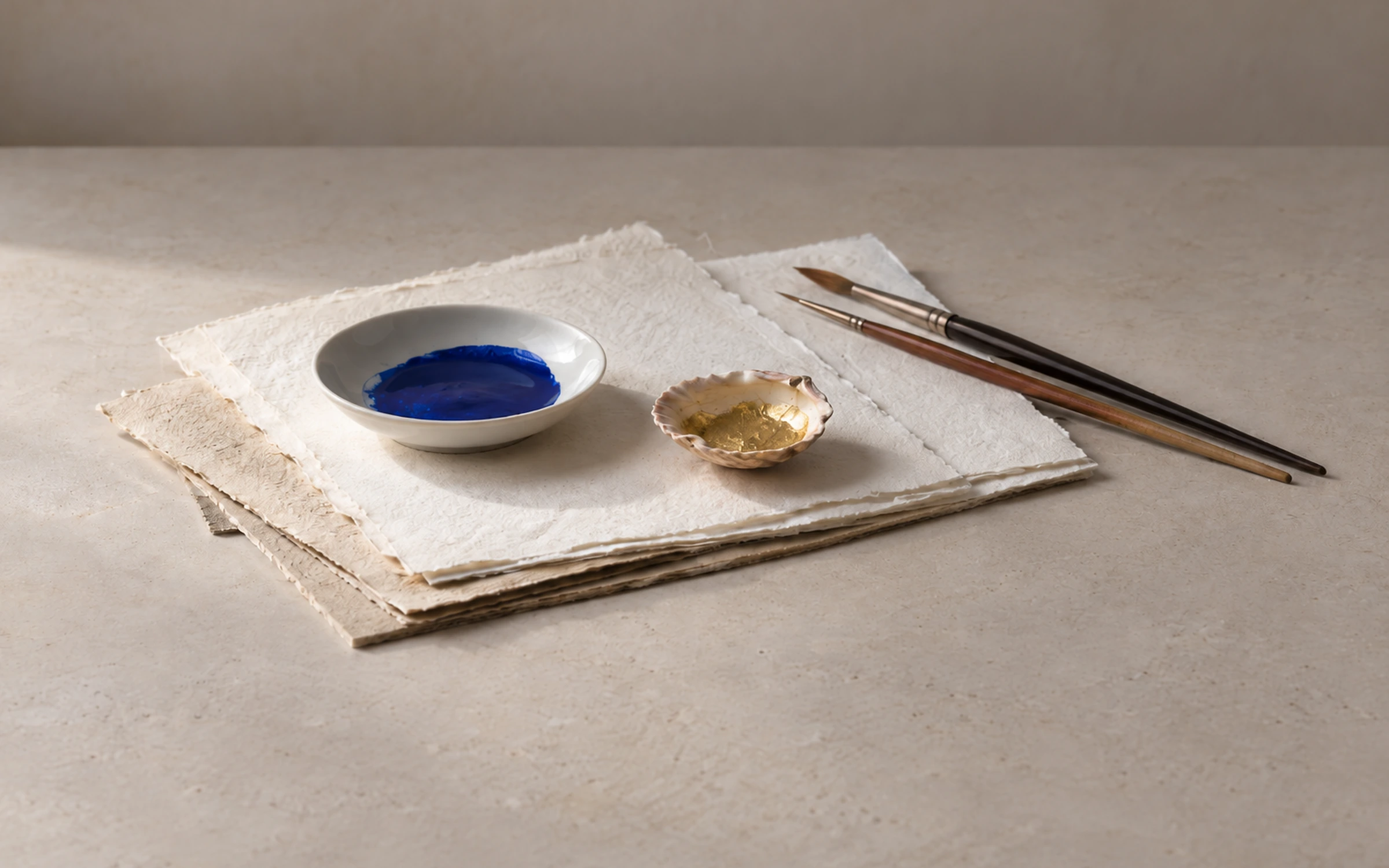 Handmade papers with ultramarine and gold pigments and fine brushes arranged on a neutral studio workbench.